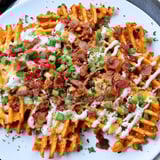 A close-up of a platter overflowing with Loaded Sweet Potato Fries, drizzled with ranch and green onions.